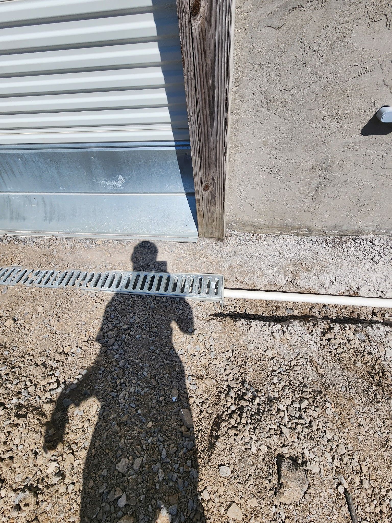 Shadow of person, grating drain, building corner, gravel ground.
