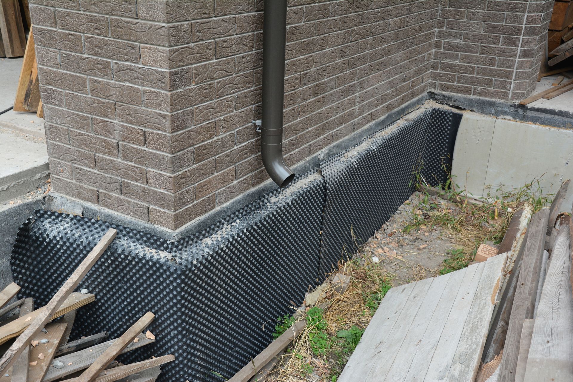 Foundation wall with black dimpled waterproofing membrane, brick, and downspout.