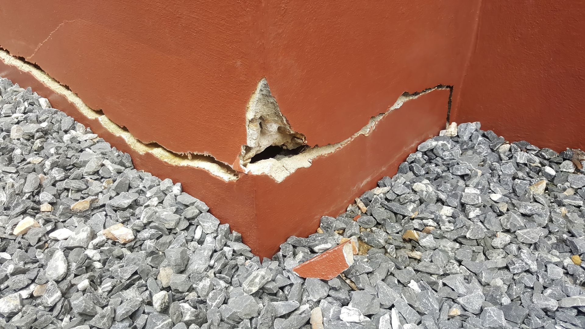 Cracked red wall with exposed damage above a bed of gray gravel