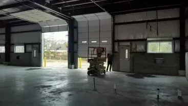 Large empty warehouse with open bay doors, concrete floor, and a person near a cart by the wall.