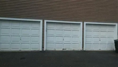Three white garage doors in a row on a brick building, with a dark driveway in front.