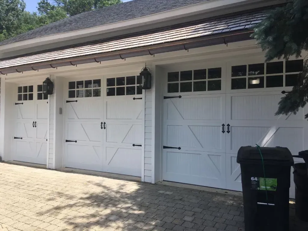 White garage doors with windows on a building exterior, beside a driveway and black trash bin.