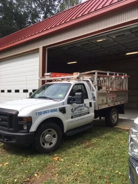 White utility truck parked beside an open garage with a red roof and metal equipment bed