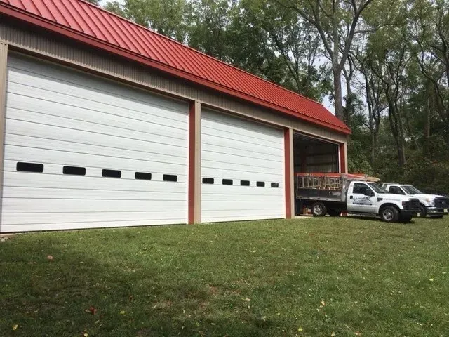 Red metal-roofed garage with two white doors, parked trucks, and a wooded yard