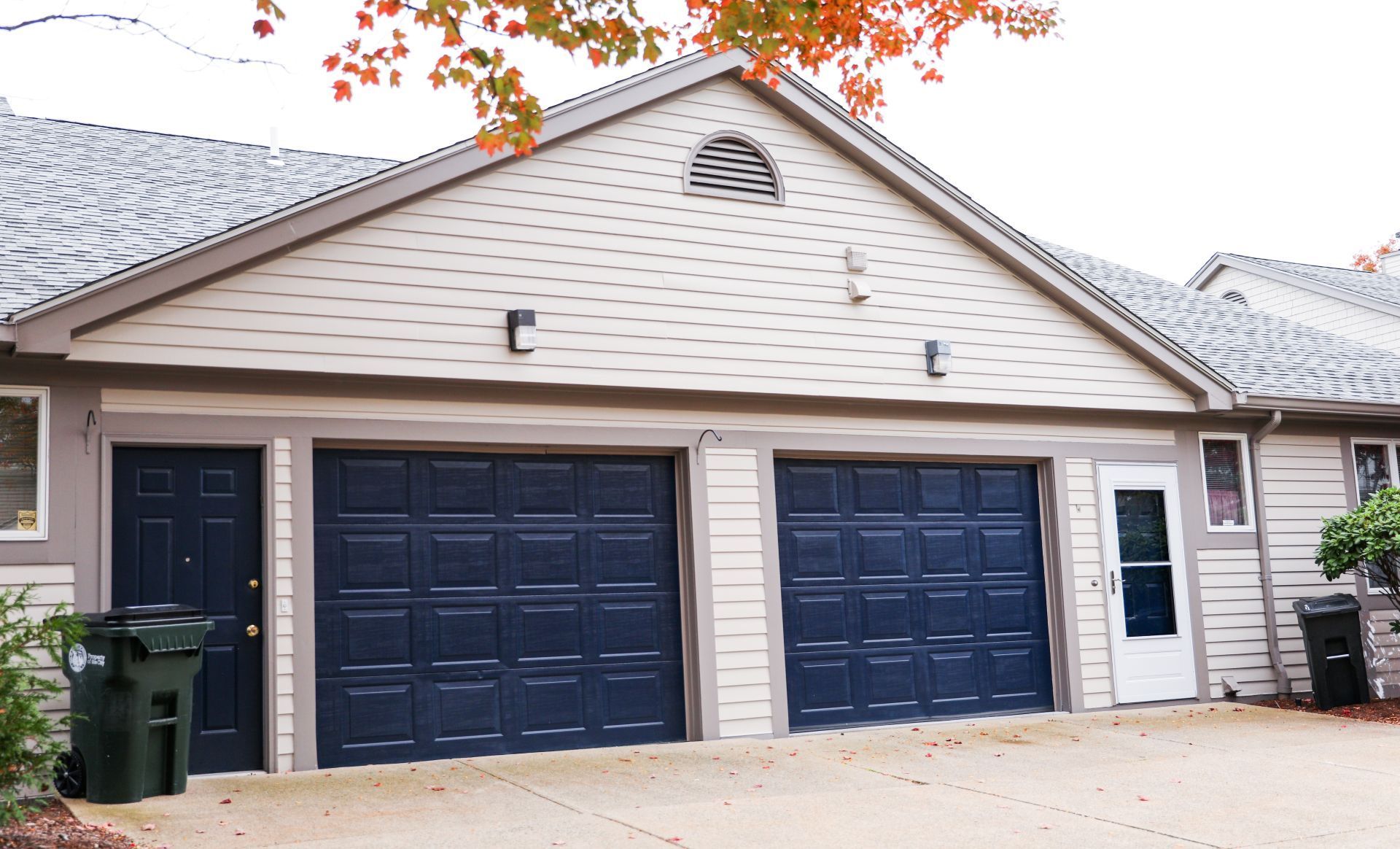 Suburban townhouse with two navy garage doors and a beige facade beneath autumn leaves