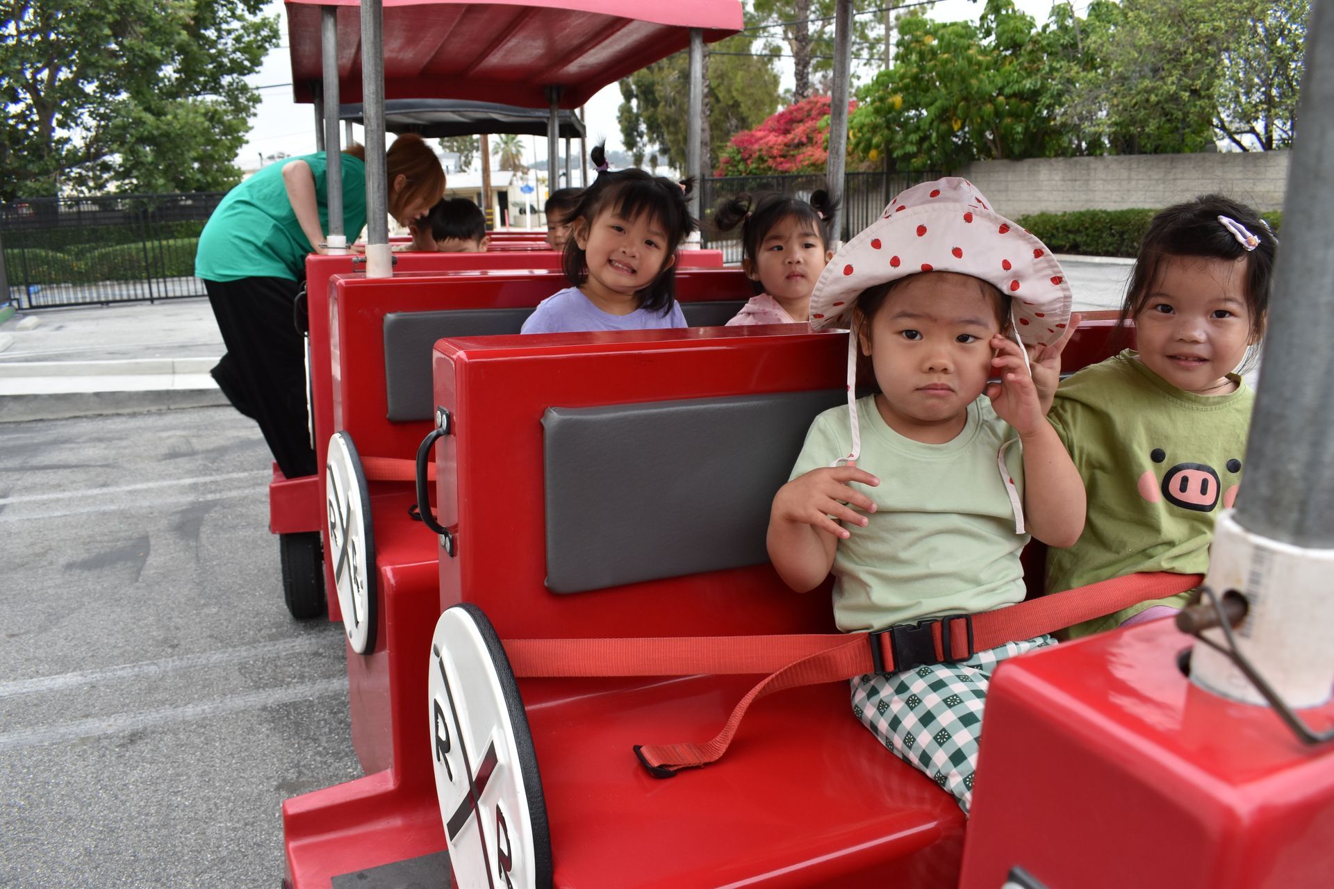 Children riding a red train, smiling. One child wearing a hat. Outdoors.