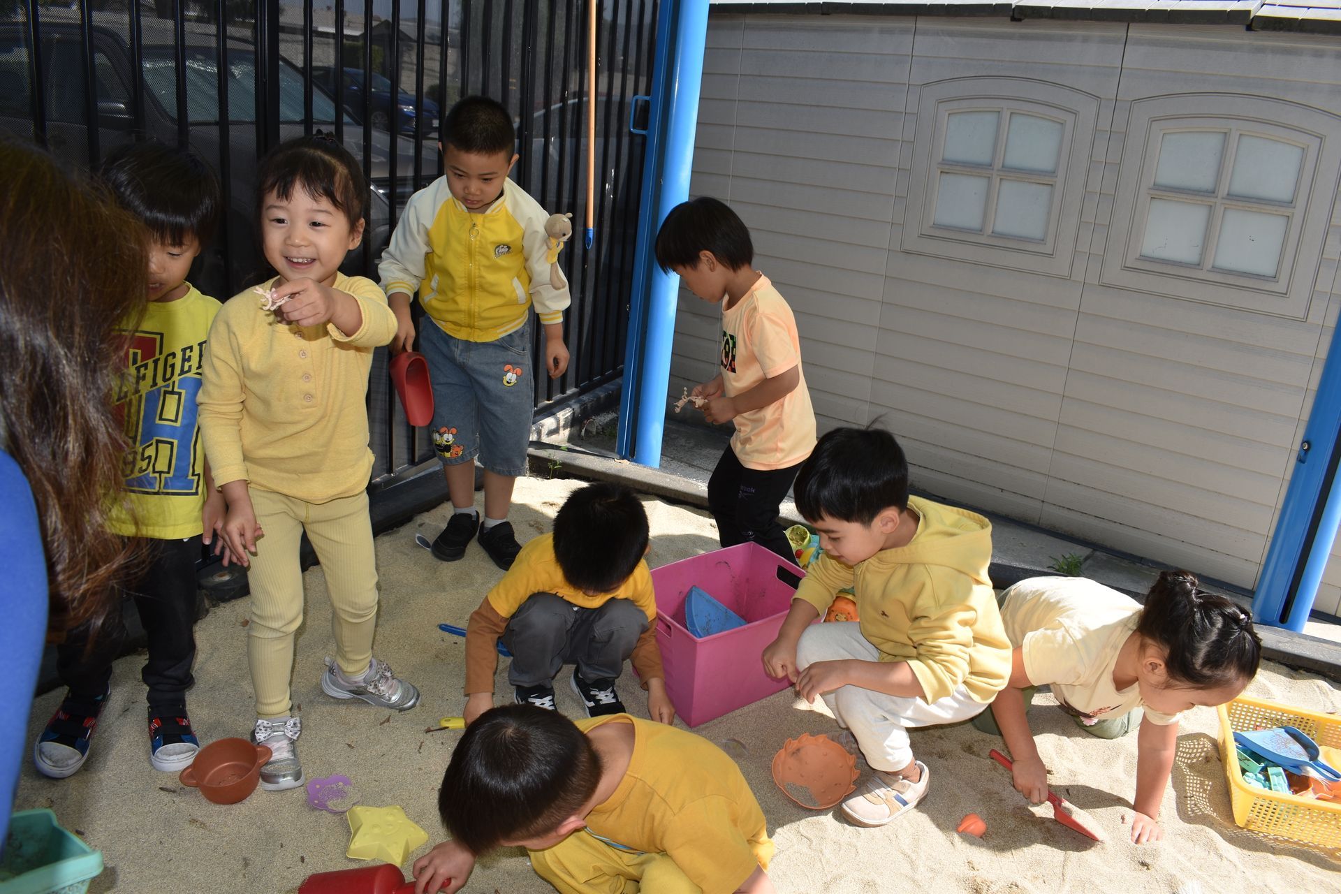 Children in yellow clothing play in a sandbox with toys, smiling.