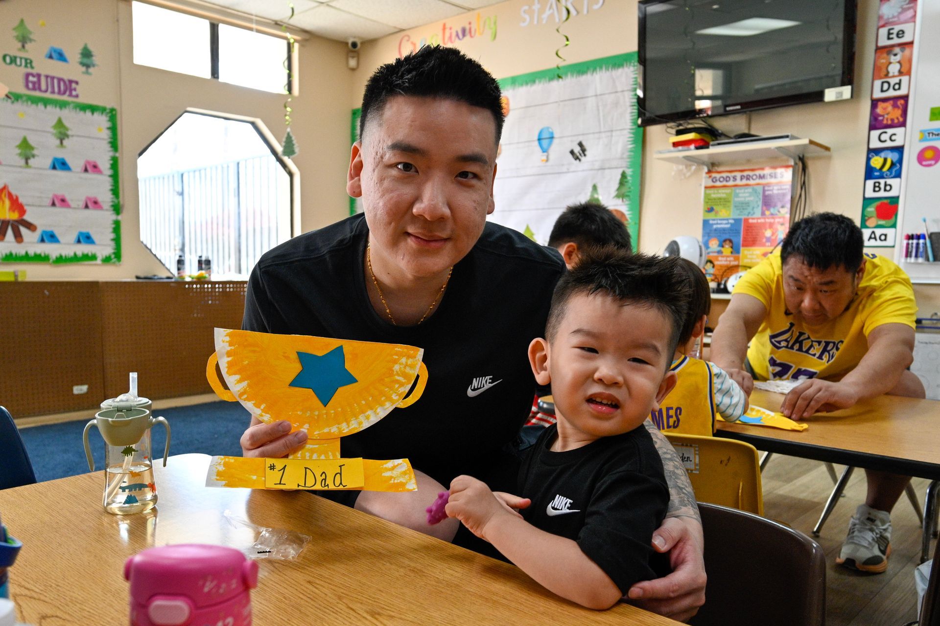 Man and child holding a paper plate trophy in a classroom. Another person in background.
