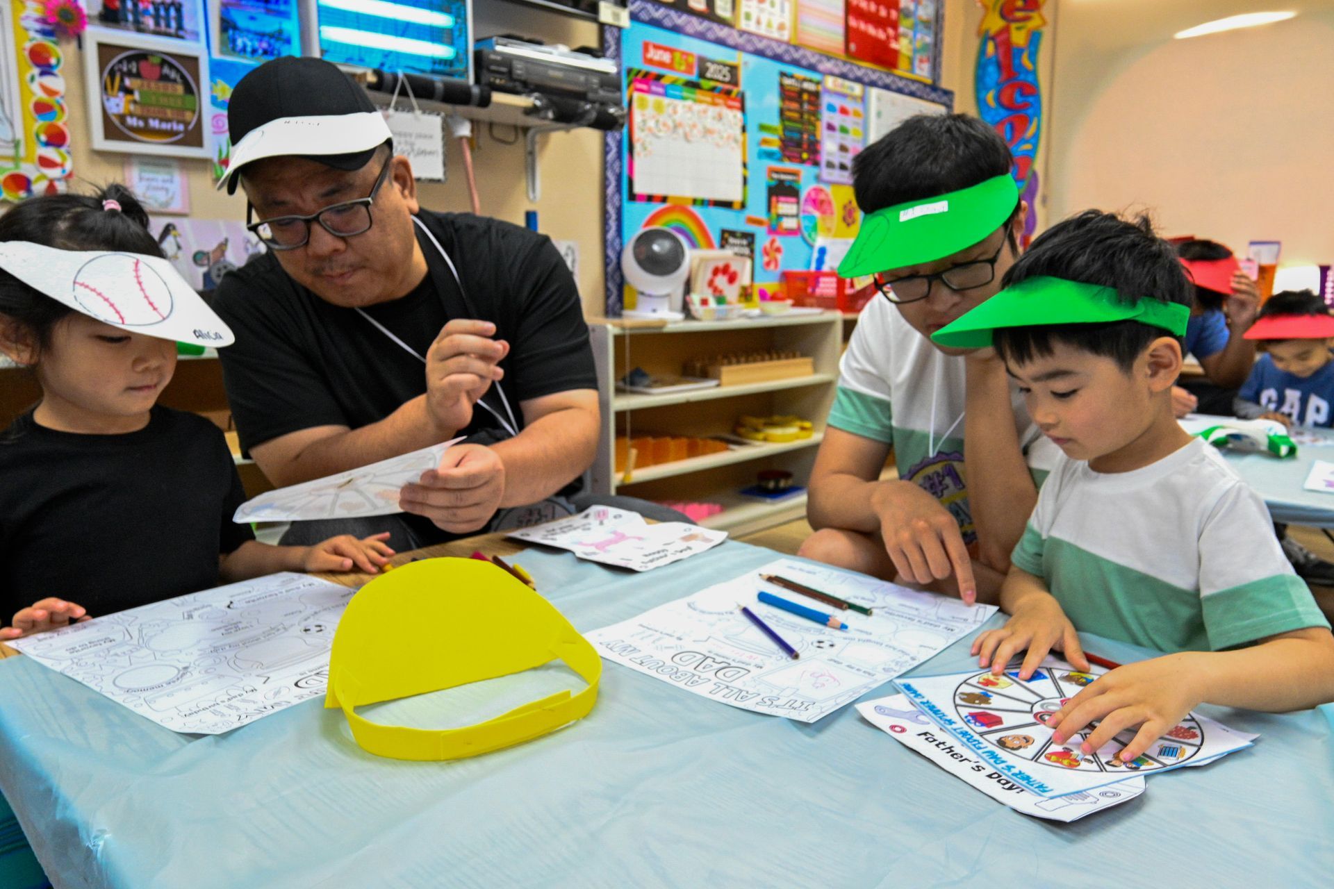 Two adults and two children at a table working on art projects in a classroom. All wearing visors.