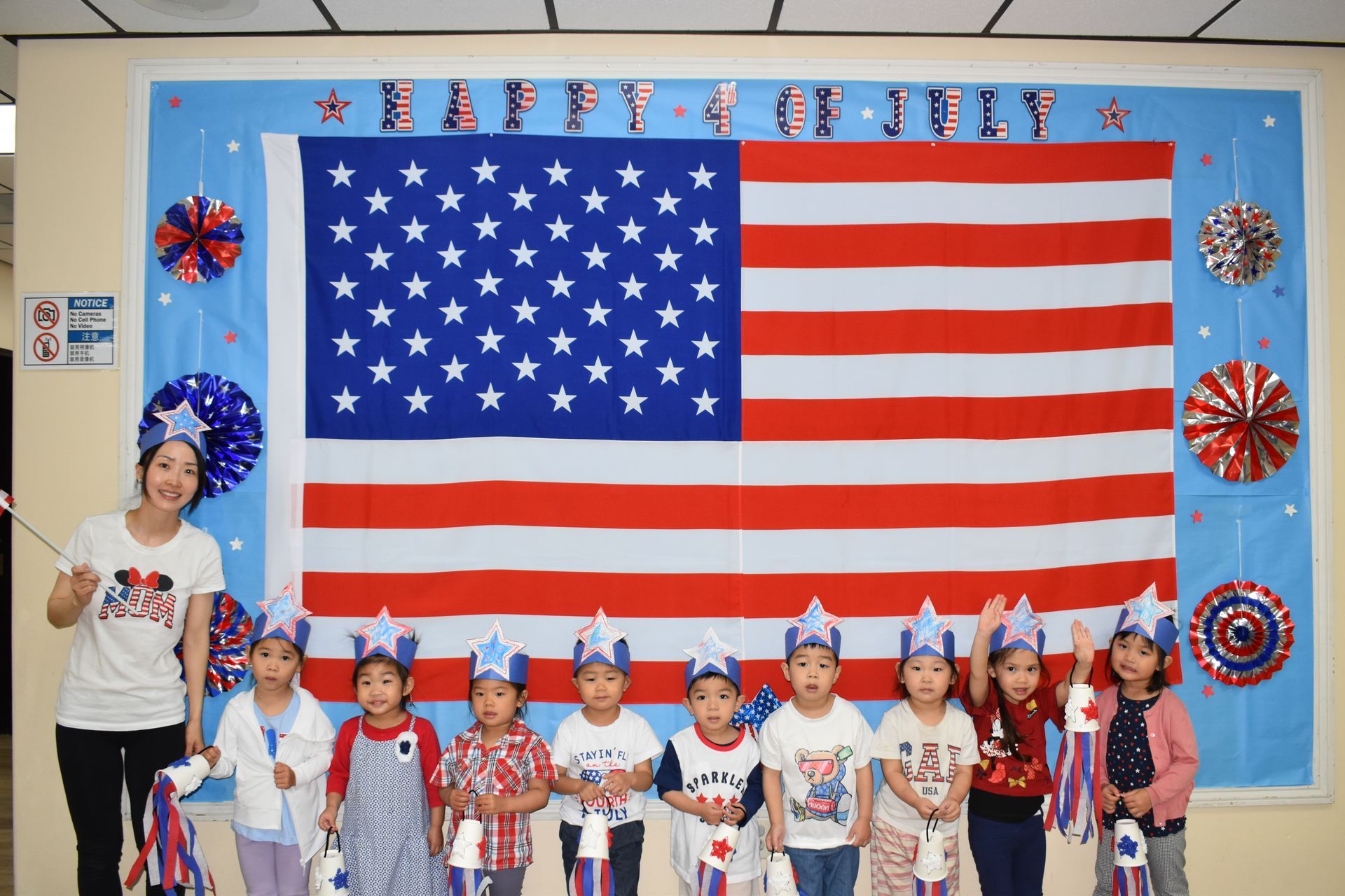Group of children and a teacher celebrate July 4th with a large American flag backdrop.
