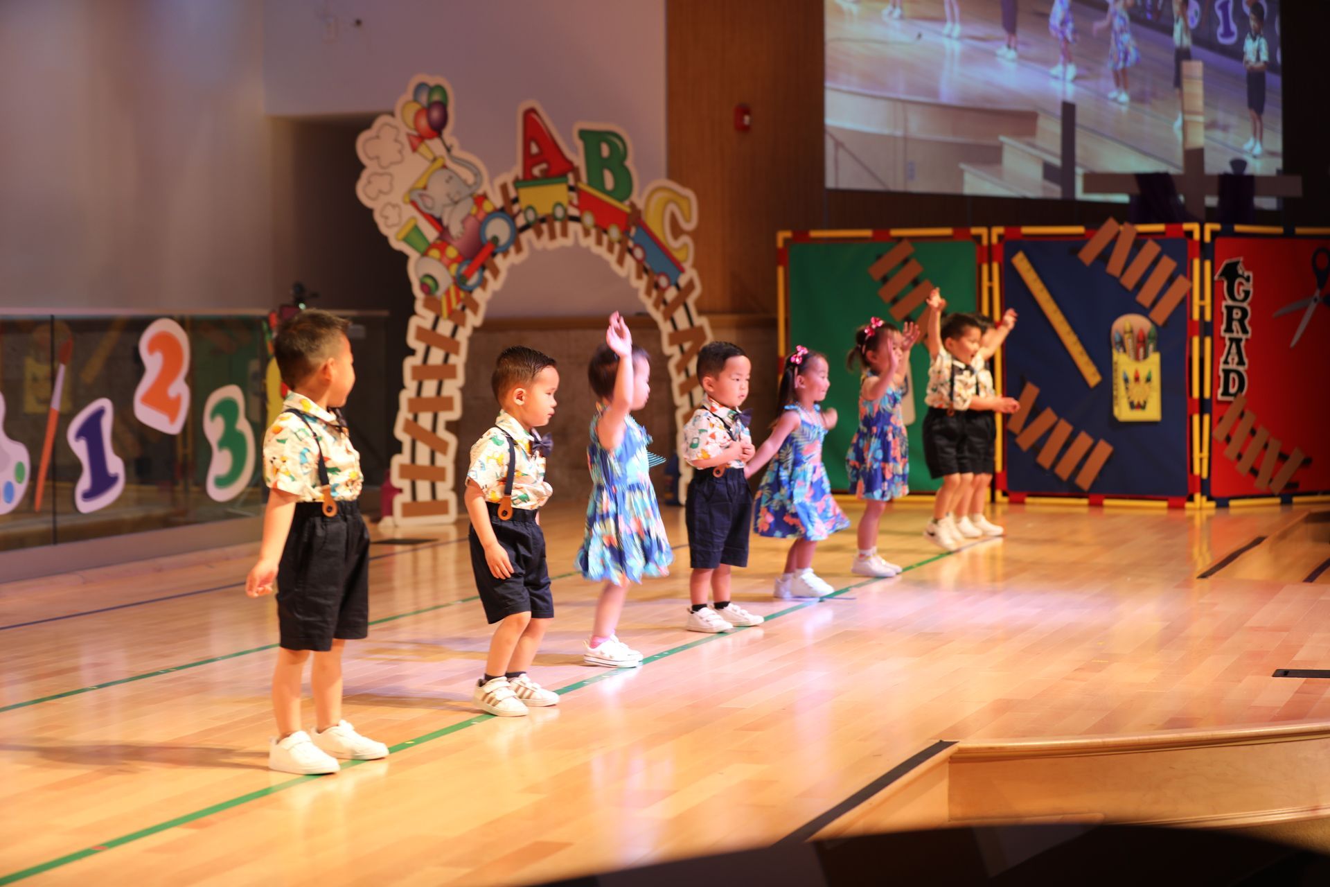 Children on a stage, performing. They wear shorts and patterned shirts, with arms raised. Archway and numbers in background.