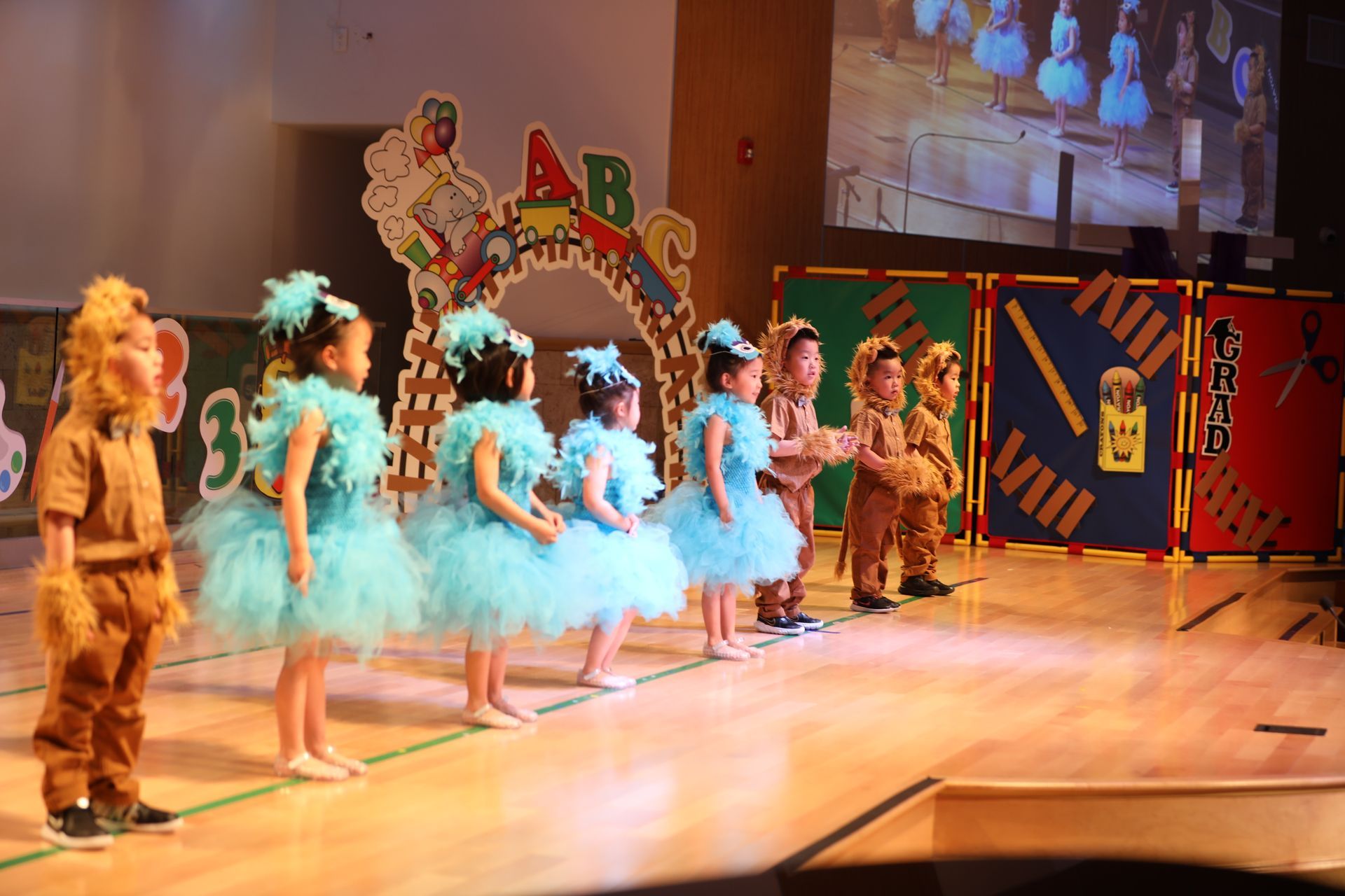 Children in costumes perform on stage, including blue tutu dresses and brown animal attire.