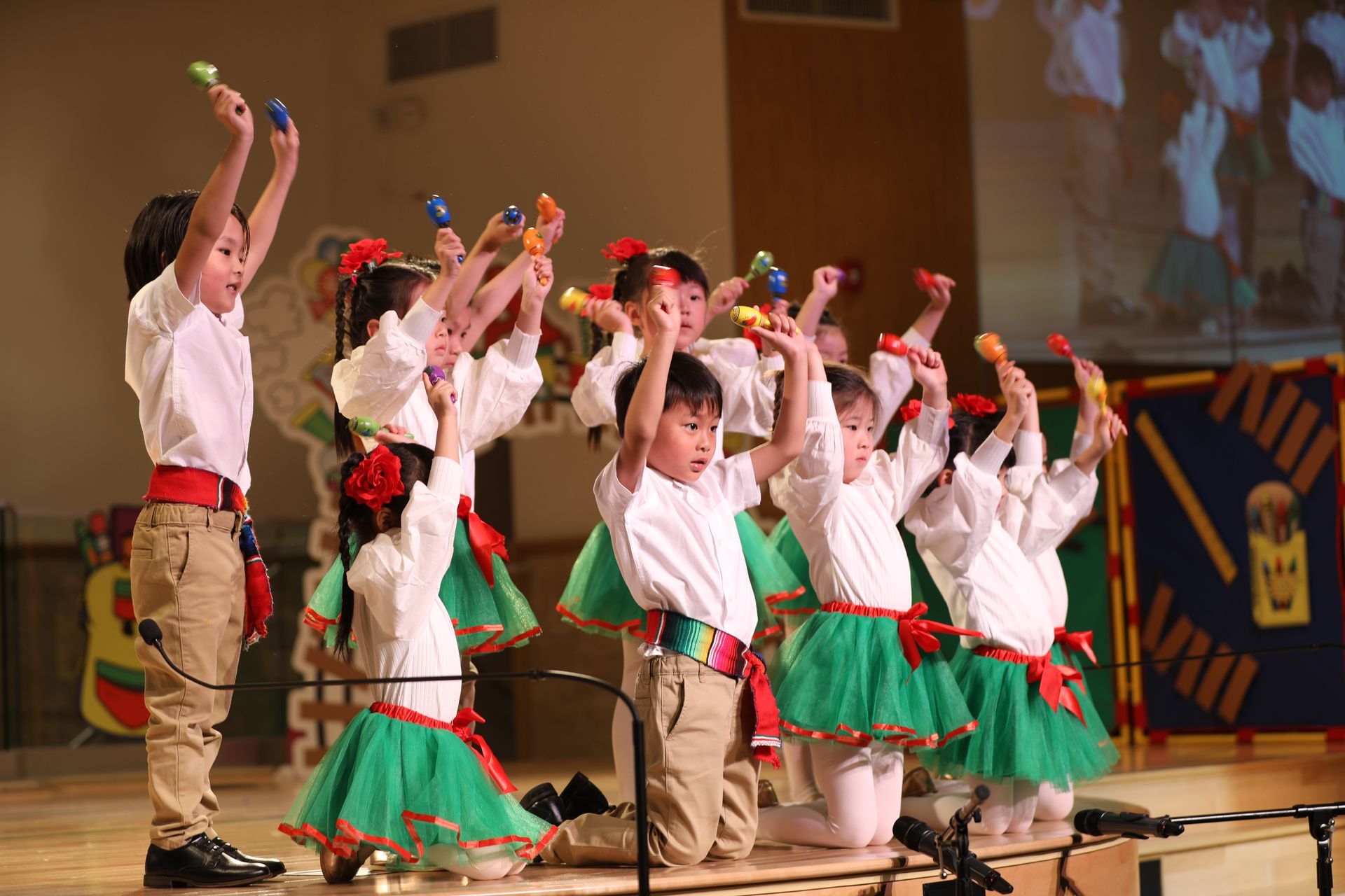Children in white shirts and green skirts, holding instruments, perform on stage.