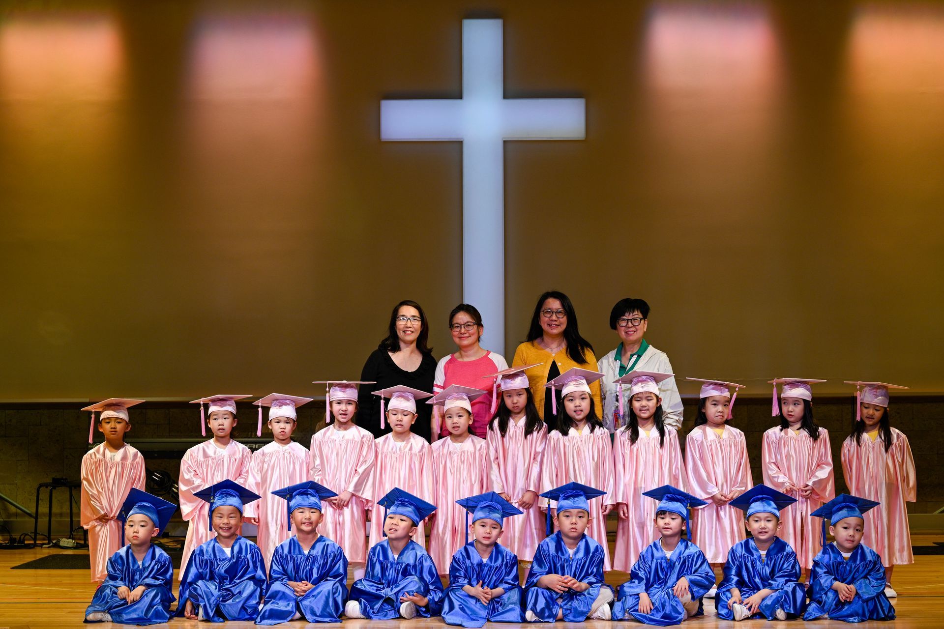 Children in graduation gowns and caps with adults in front of a cross on a stage.