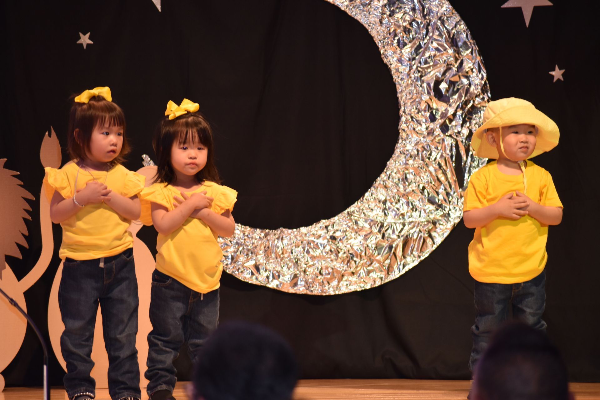 Children in yellow shirts and hats on stage with a moon backdrop, holding hands over their hearts.
