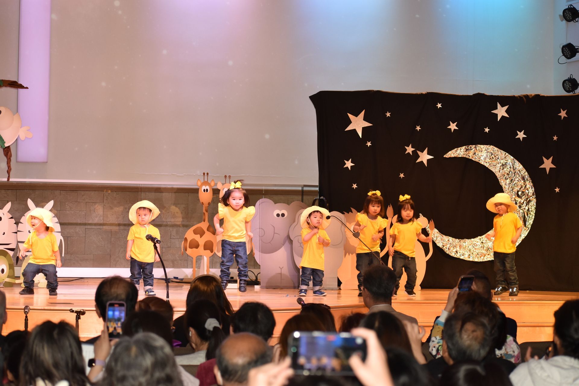 Children in yellow shirts perform on stage, with a crescent moon backdrop, audience watching.