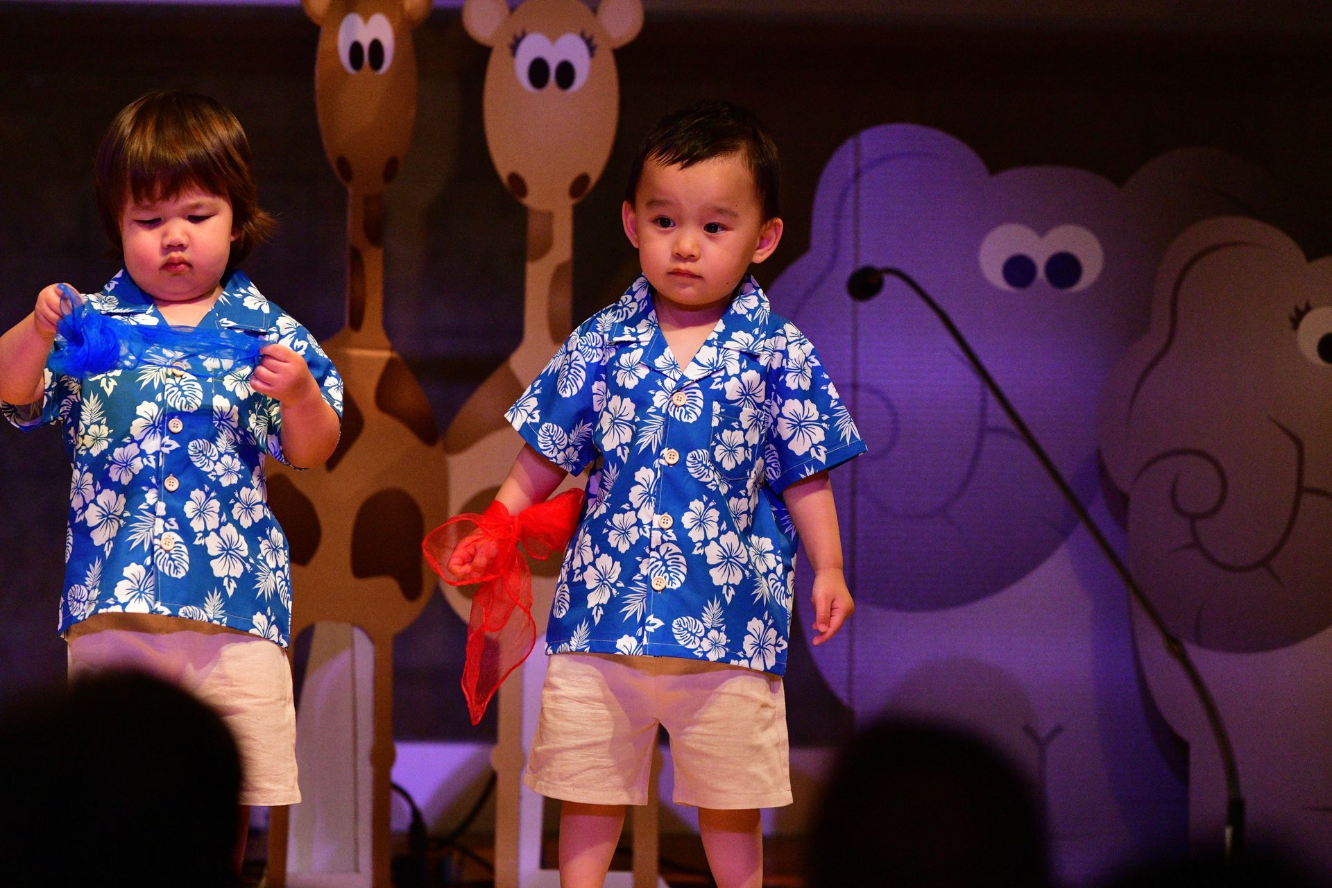 Two young children in blue floral shirts, tan shorts on stage, holding scarves, with animal cutouts.