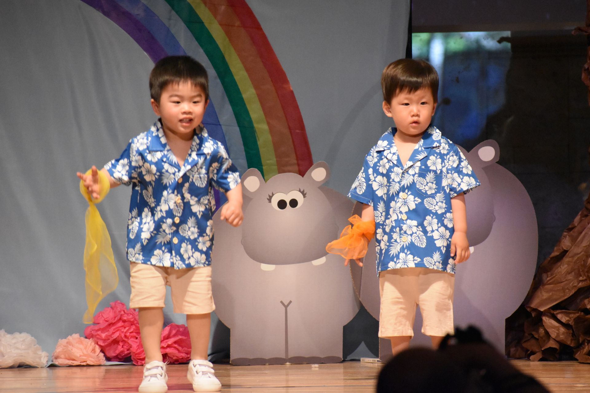 Two young children wearing matching blue shirts and khaki shorts perform on stage.
