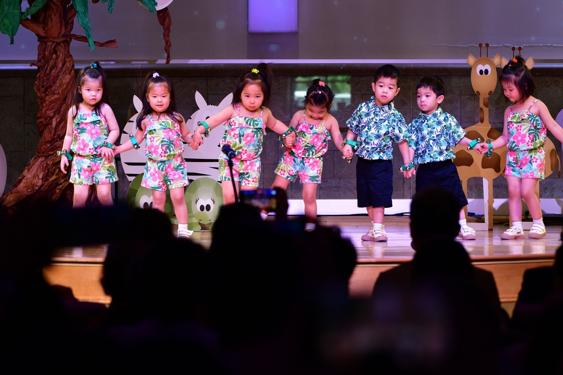 Children in floral outfits on stage, holding hands, performing. Audience in foreground.
