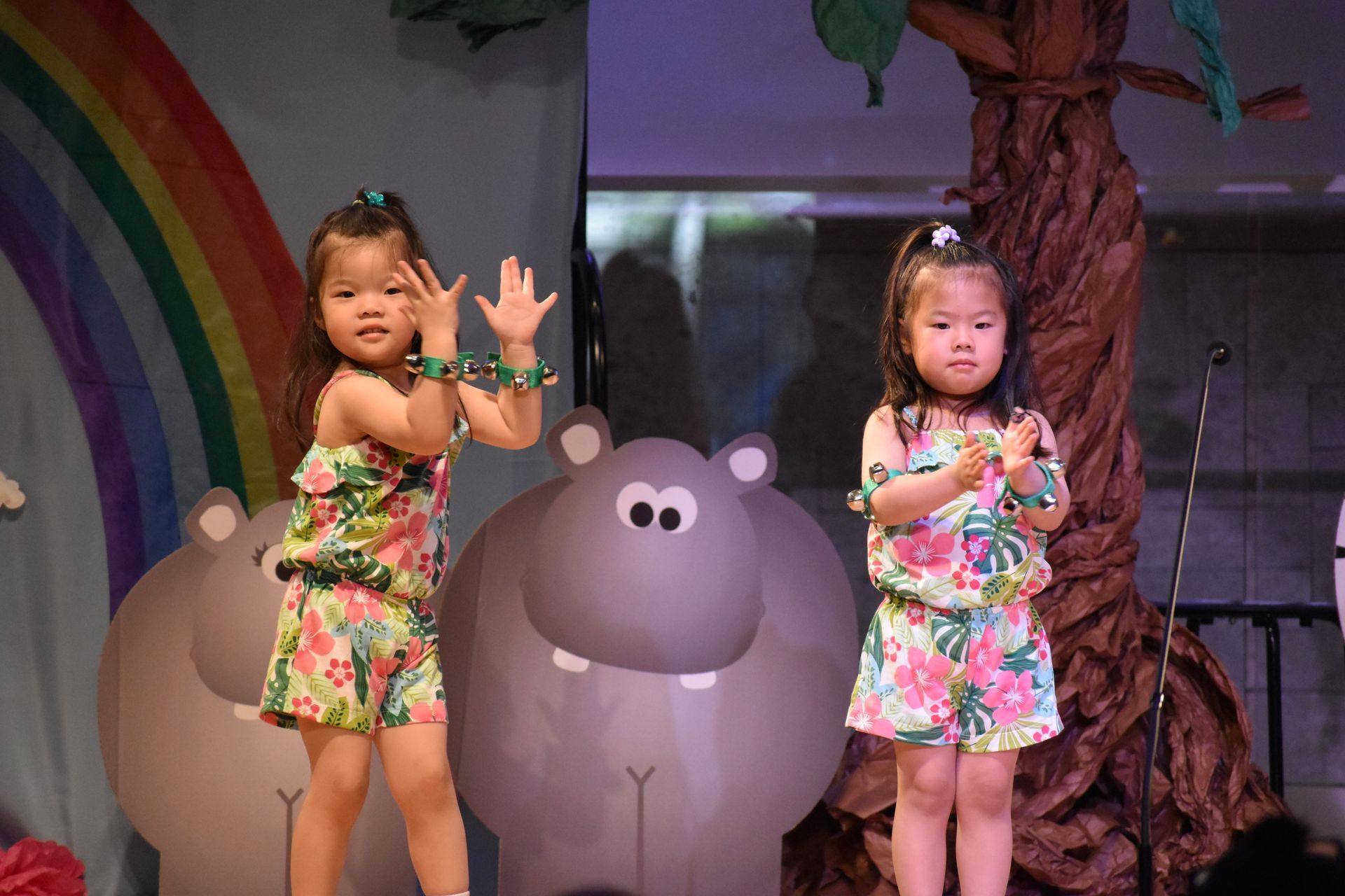Two young children in floral outfits dance on stage with a hippo and rainbow backdrop.