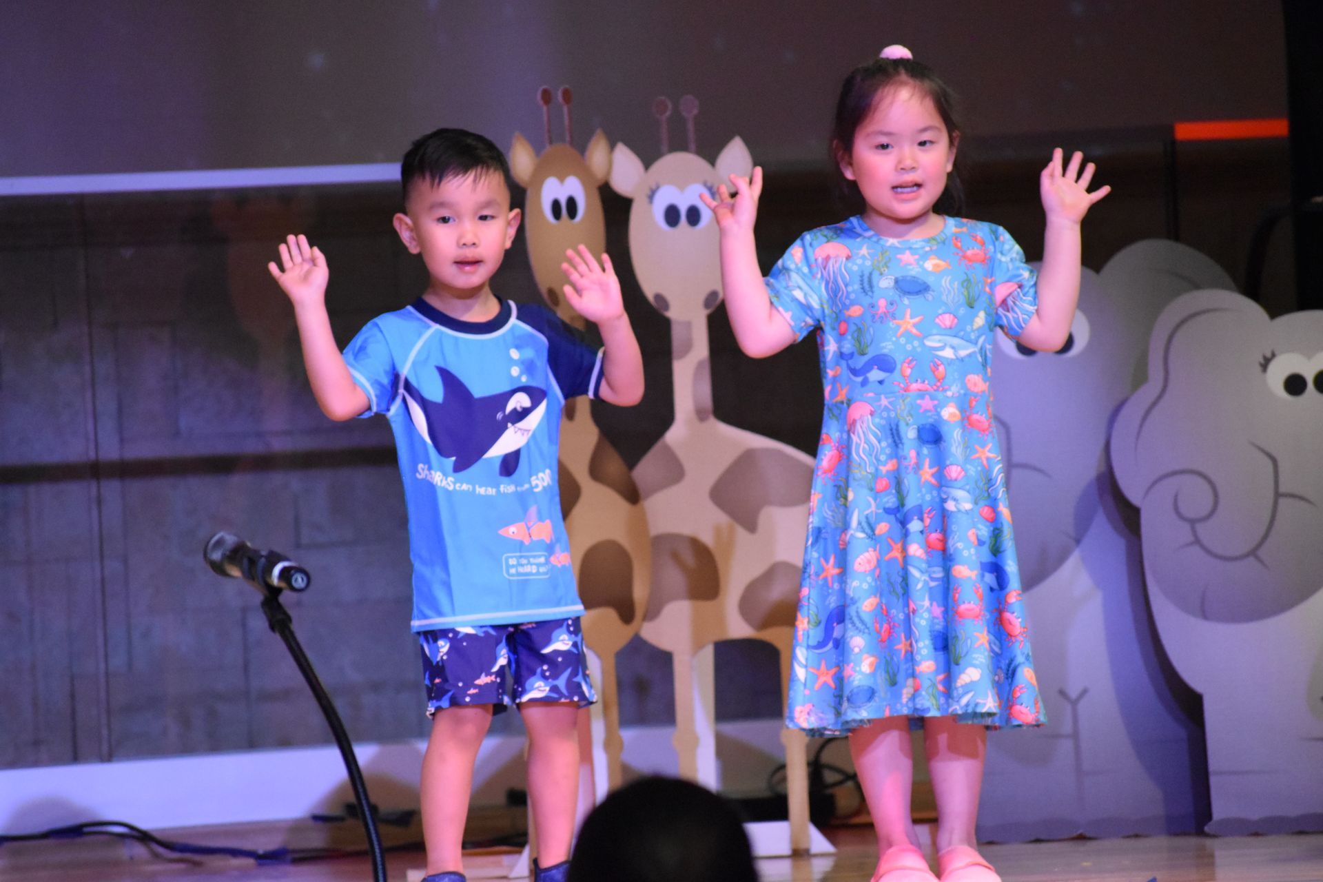 Two children on stage with hands raised, smiling. Behind them, animal cutouts.