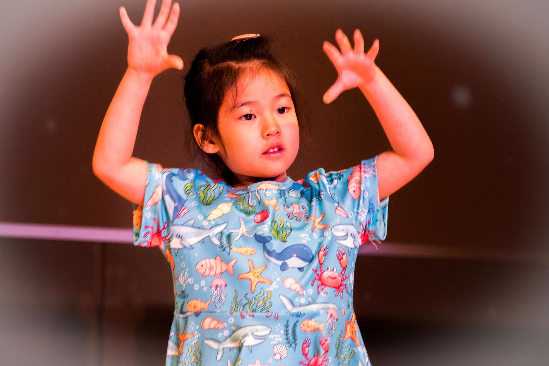 Girl with hands raised, wearing a blue ocean-themed dress, performing on stage.
