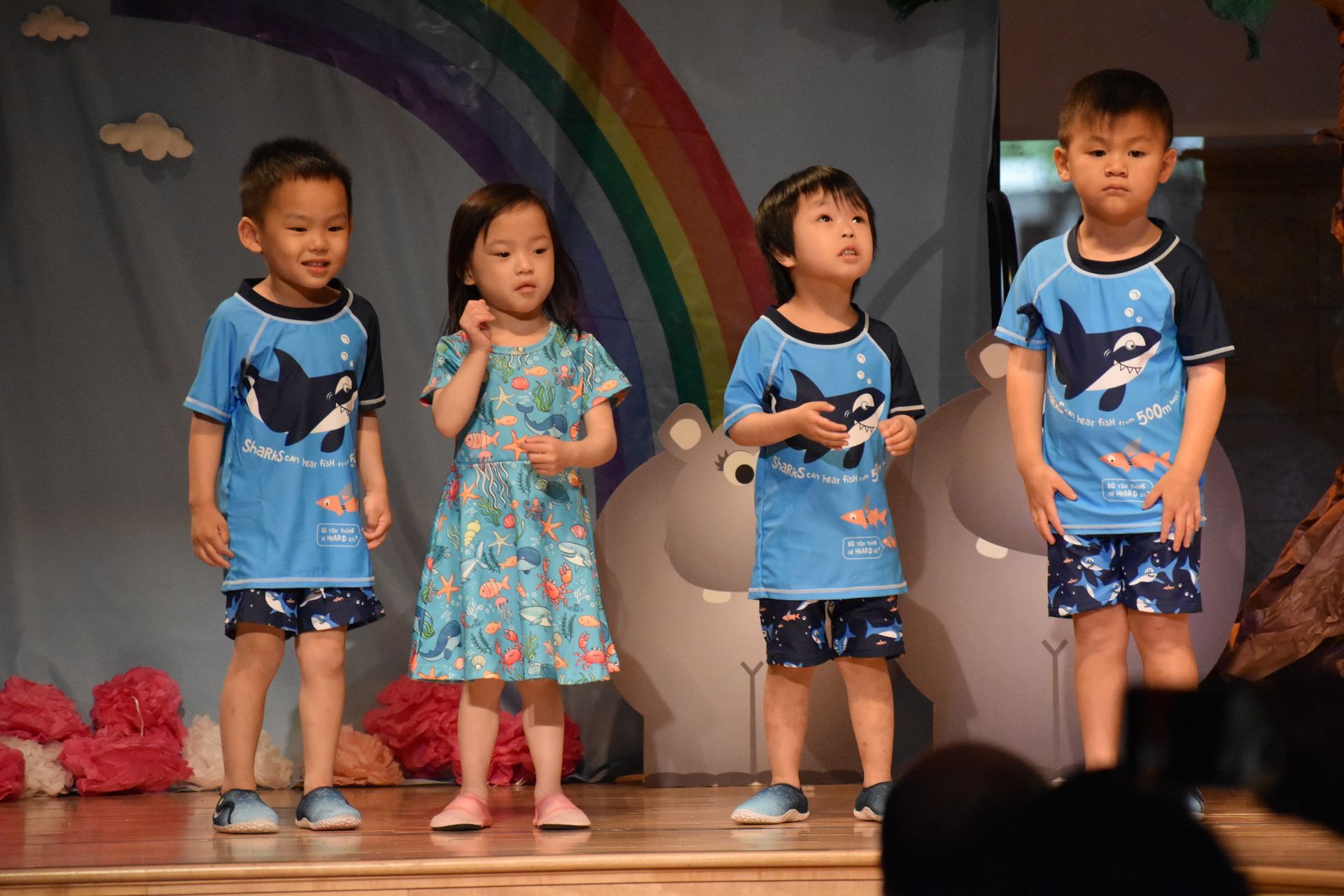 Children in swimsuits on a stage with a rainbow backdrop. They are performing a song or presentation.