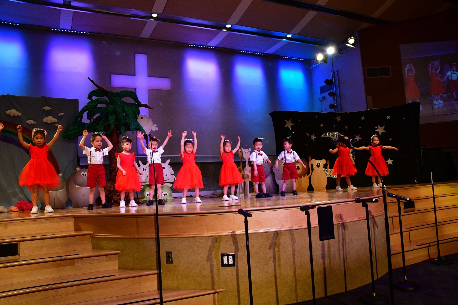 Children performing on a stage with a cross backdrop; some in red dresses, others in white shirts and red shorts.
