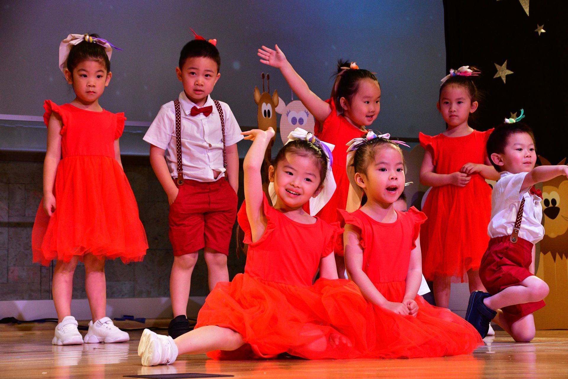 Children in red dresses and shorts on a stage, some dancing with smiles.
