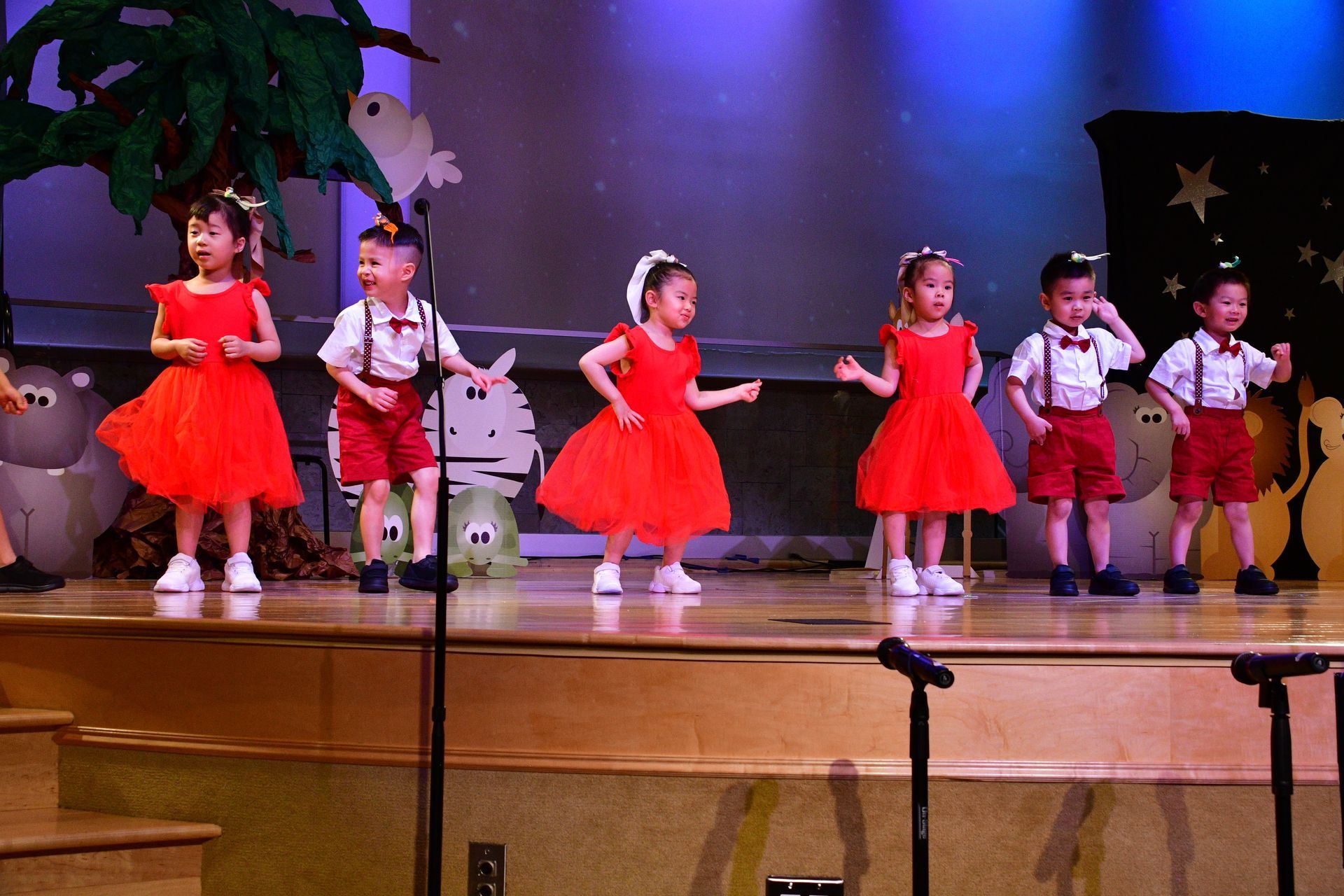 Children in red outfits performing on a stage with a backdrop of stars and trees.