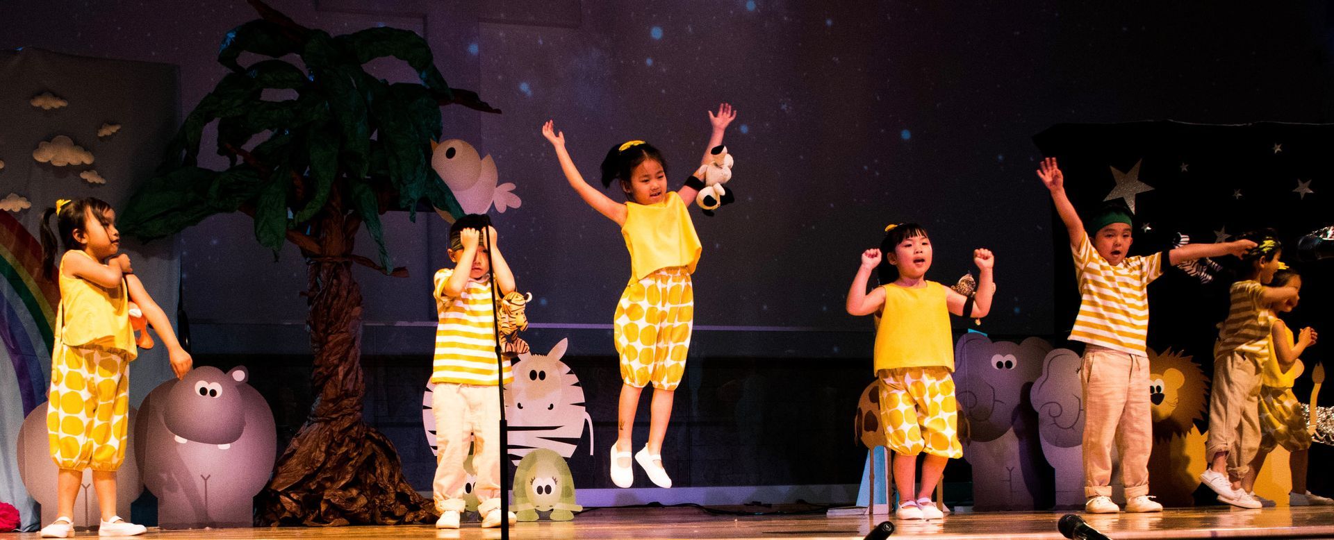 Children performing on stage, wearing yellow, against a backdrop with a tree and animal figures.