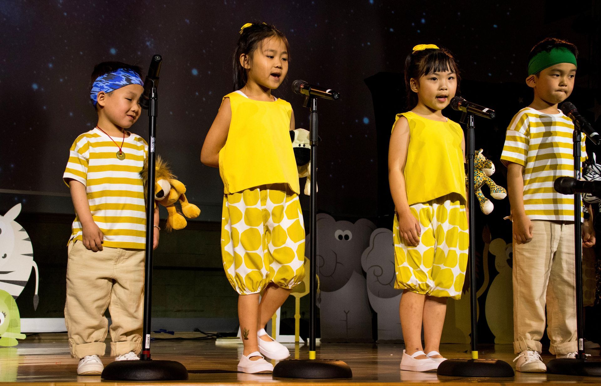 Four children on stage singing, wearing yellow tops and pants. Holding microphones.