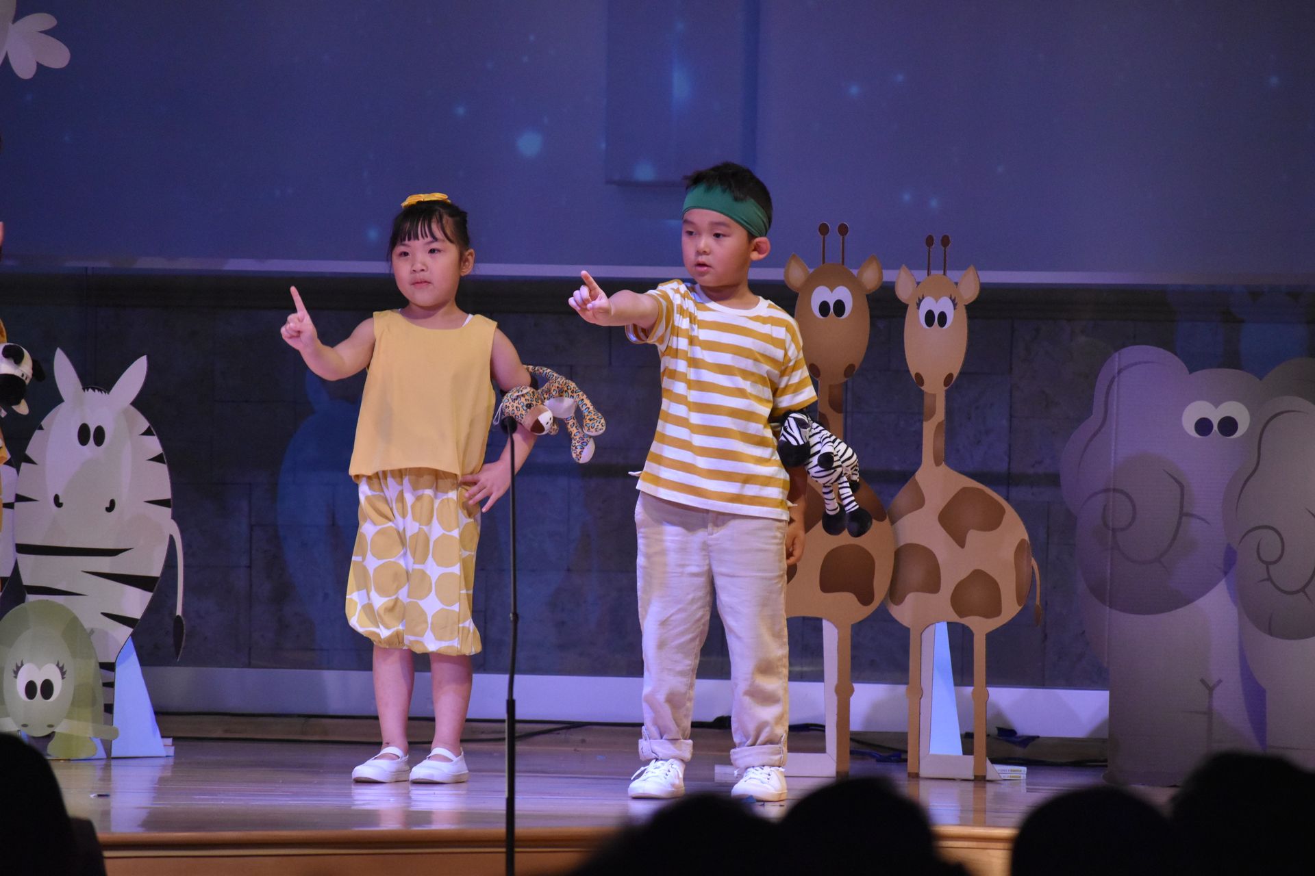 Two children in yellow outfits on stage, pointing, with animal cutouts behind them.