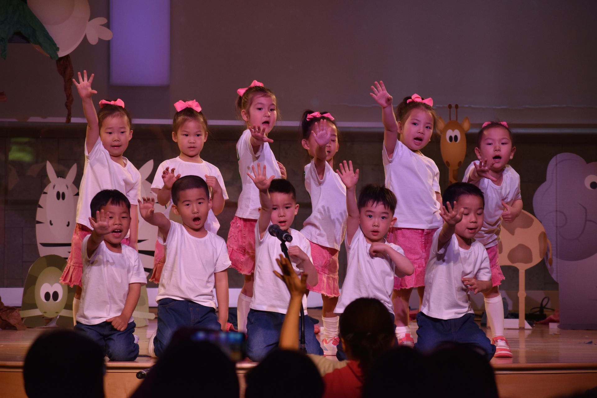 Children on stage in white shirts and pink bows, waving to the audience. Animal cutouts decorate the backdrop.
