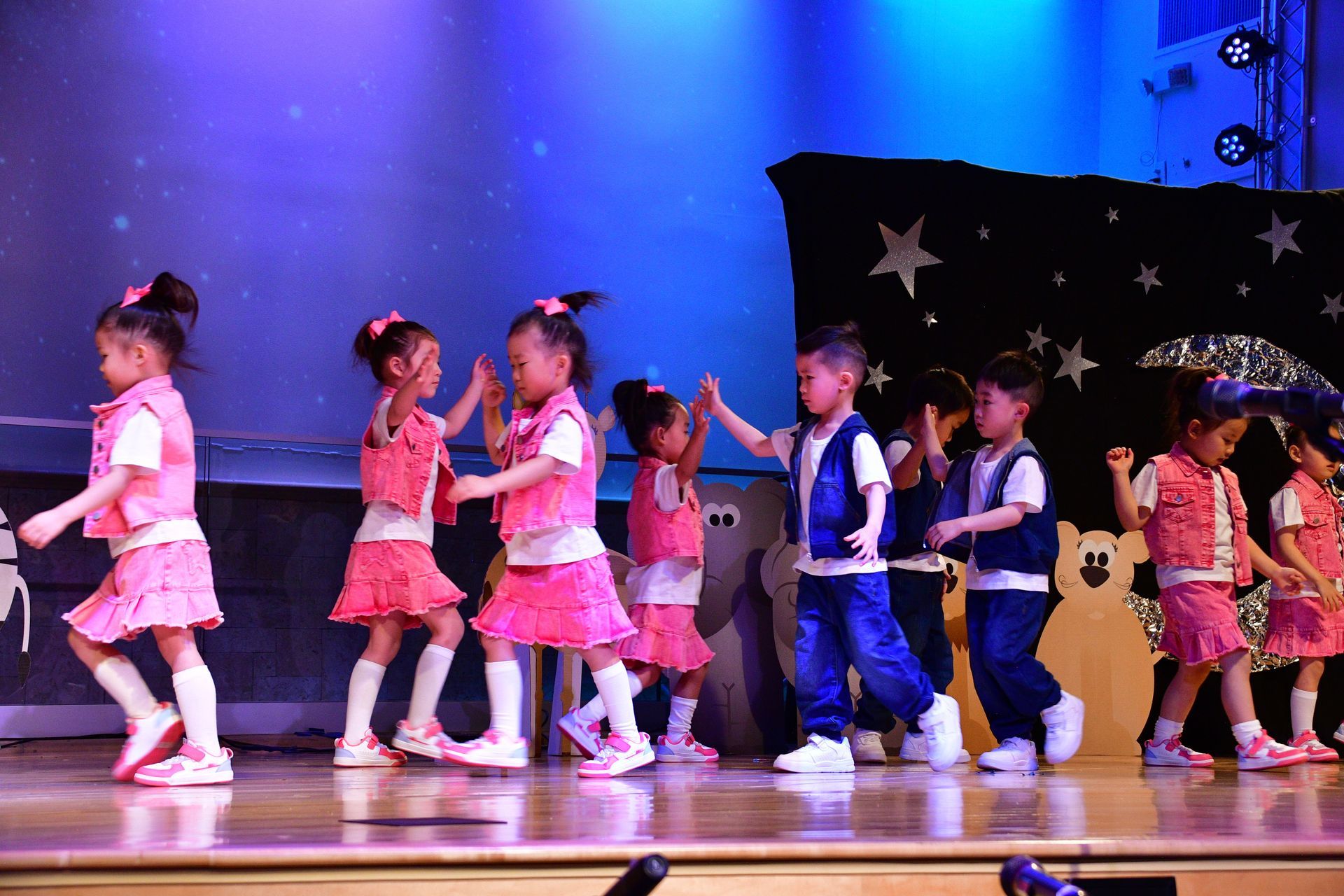 Children in pink and blue outfits dancing on stage under stage lighting.