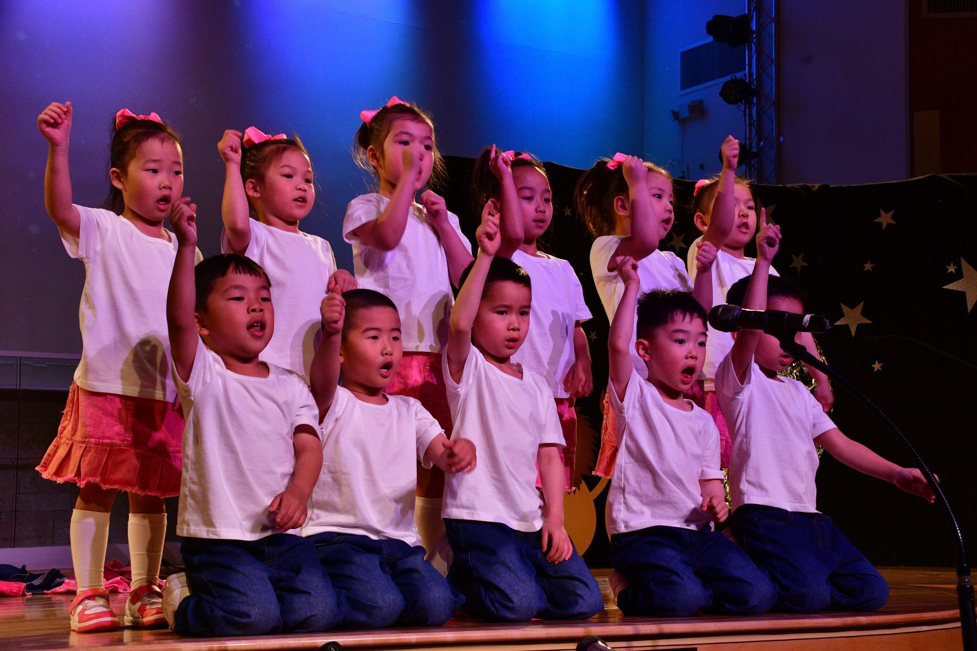 Children in white shirts and jeans on a stage, singing with arms raised under blue stage lights.