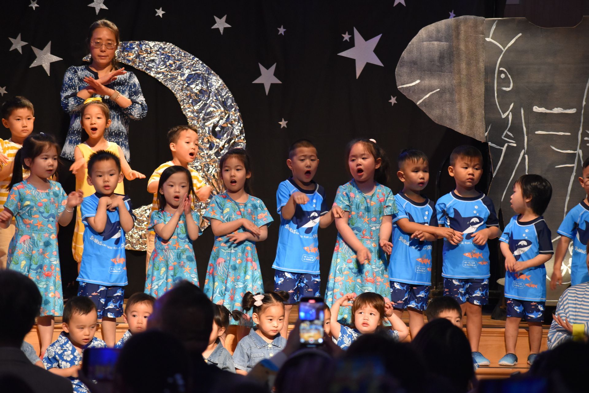 Children in blue outfits sing on stage with an adult. Black backdrop with stars and moon.