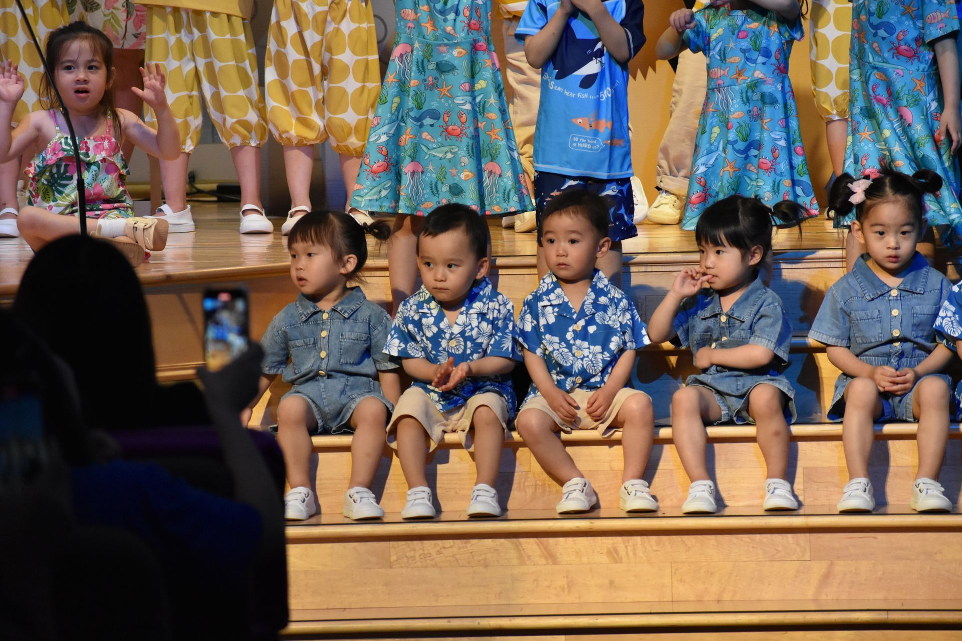 Children in matching blue outfits on stage, some with floral shirts, posing for a photo.