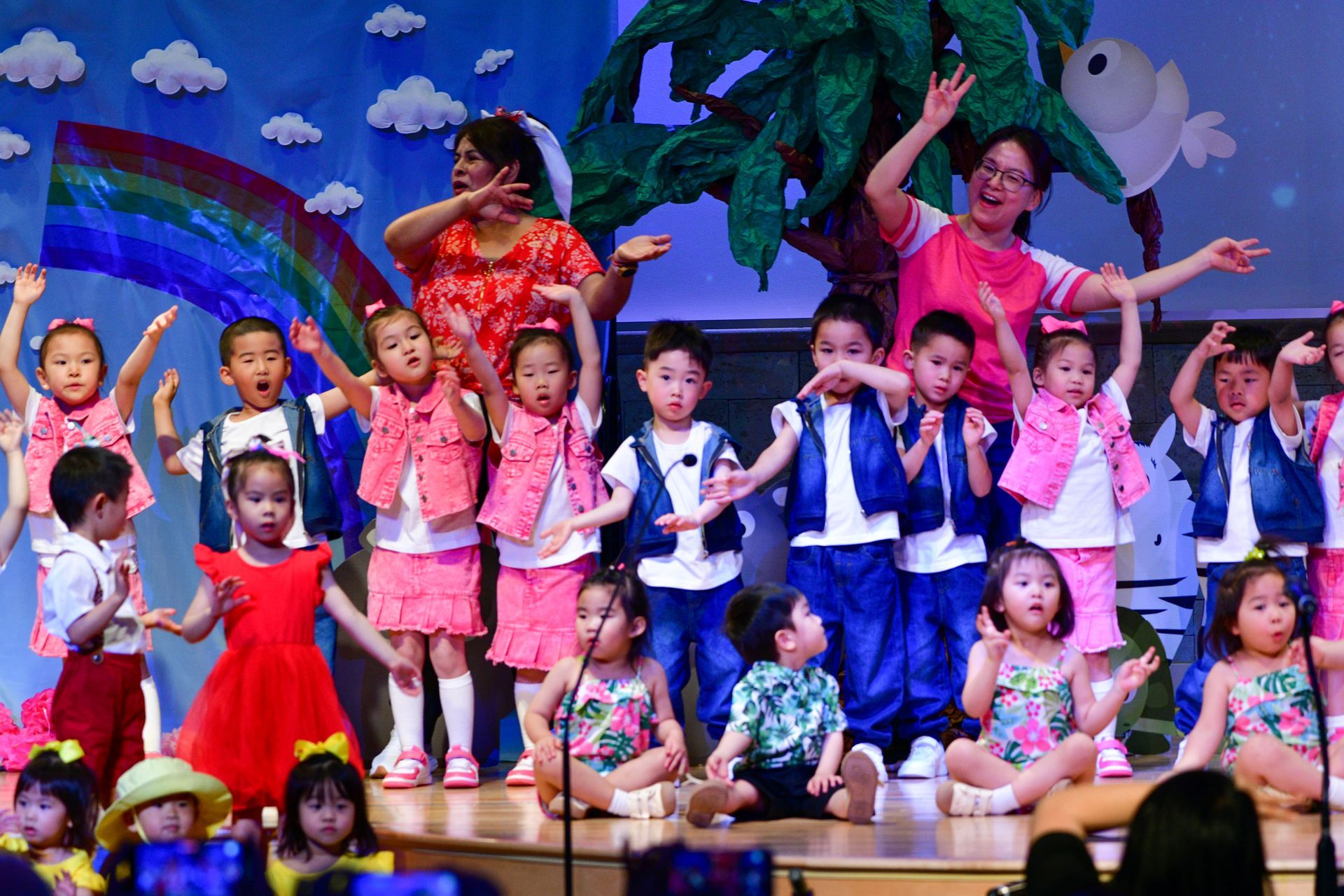 Children in performance outfits on stage with two adults. Rainbow and tree backdrop.