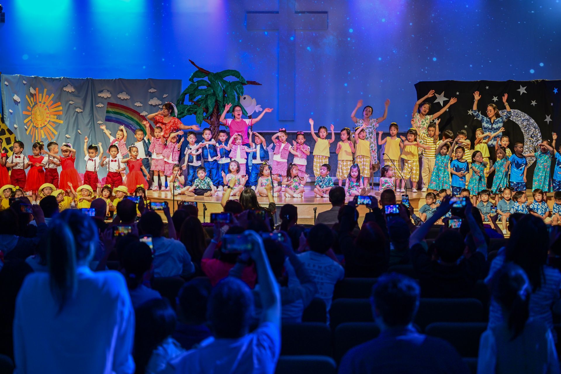 Children in costumes perform on a stage with a backdrop of a painted sun, rainbow, and stars; audience watches.