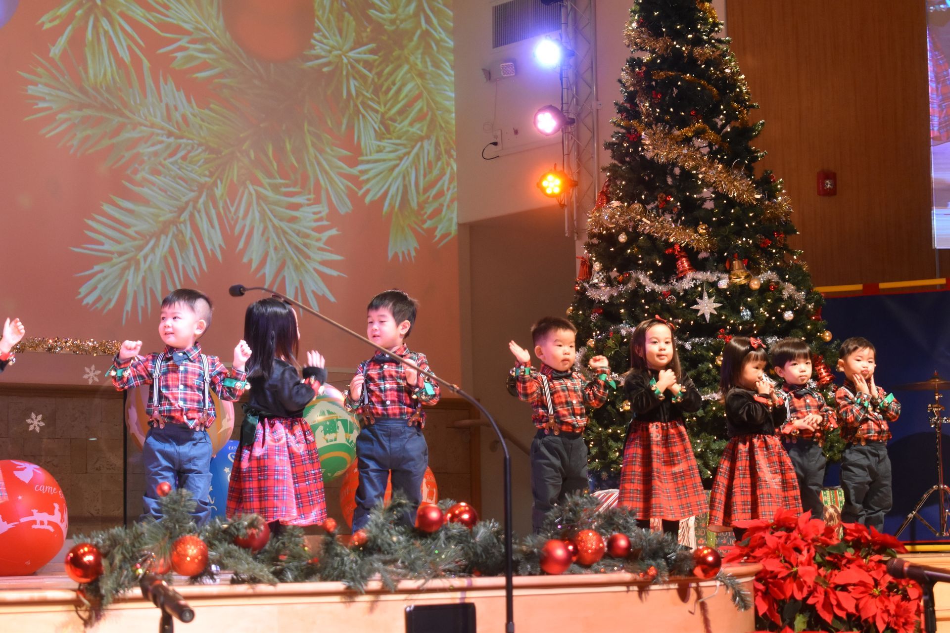Children on stage in holiday outfits performing by a decorated Christmas tree.