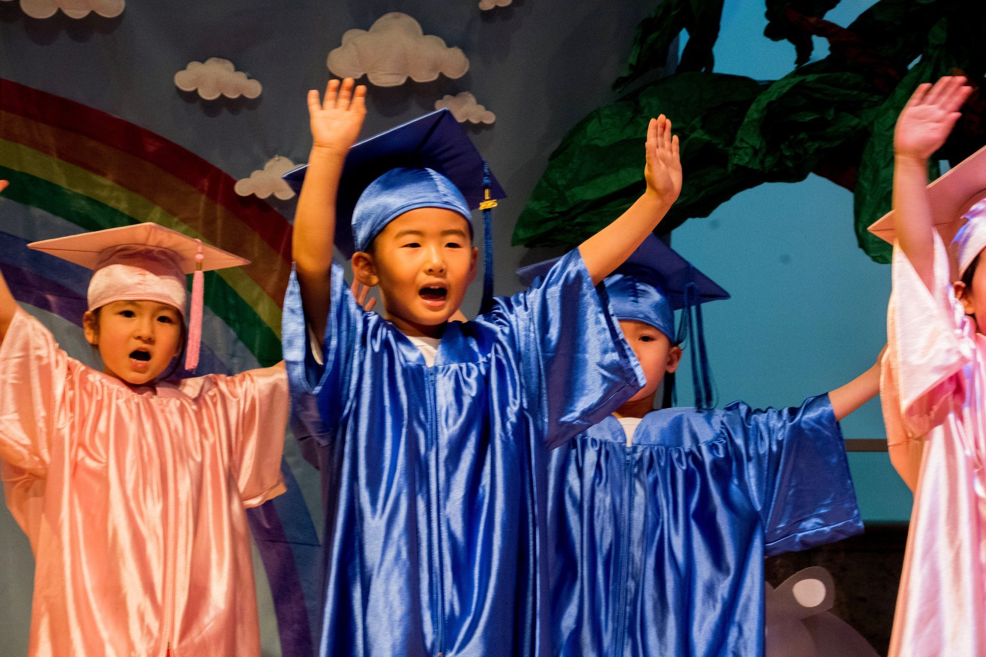Children in graduation gowns and caps raise their hands on stage.