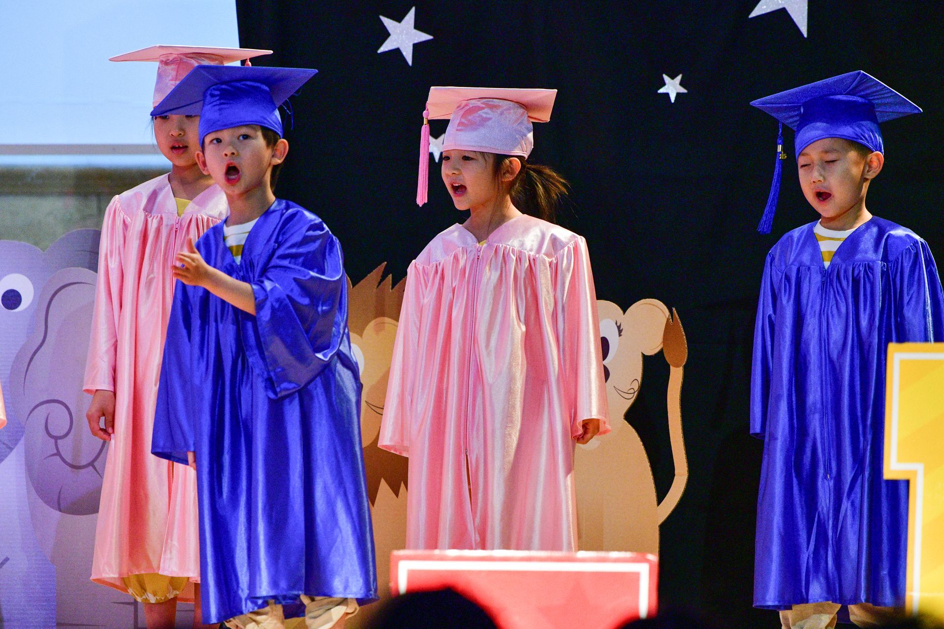Children in graduation gowns singing on a stage decorated with animal cutouts and stars.