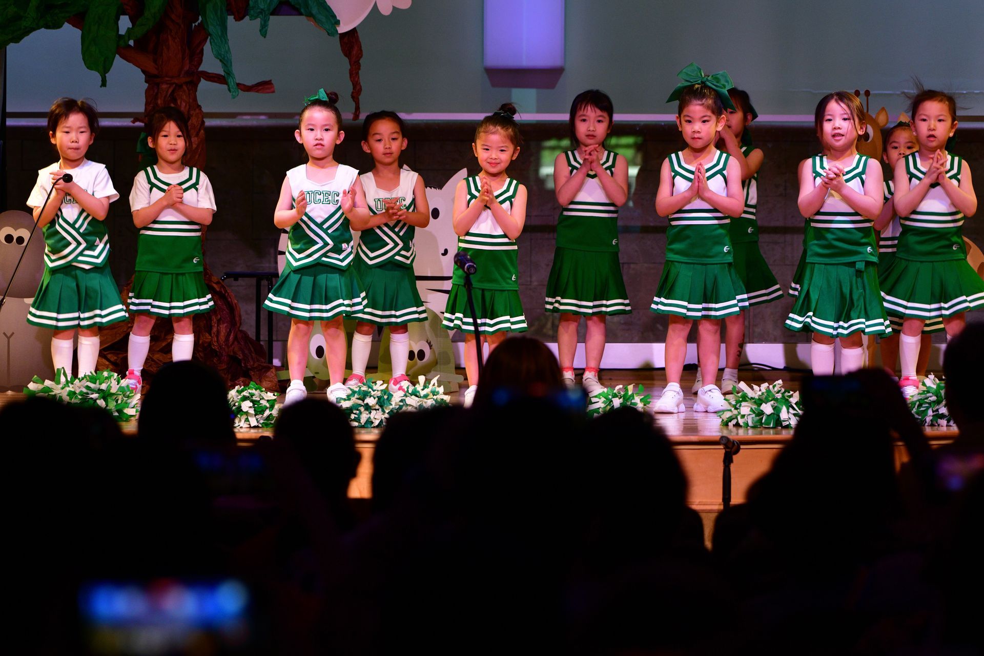 Children in green cheerleading outfits performing on stage, hands clasped.