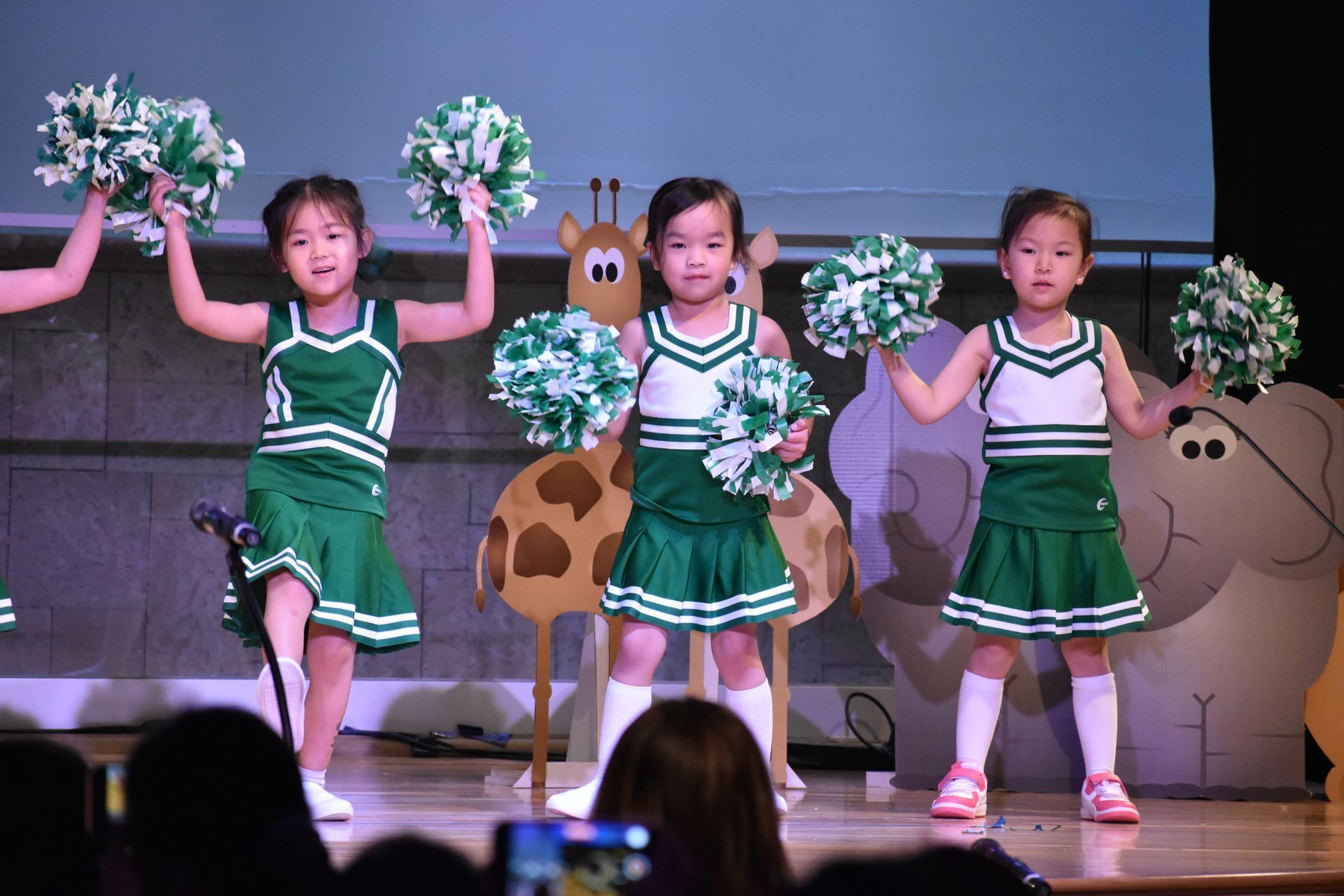 Three young cheerleaders in green and white uniforms perform on stage with pom-poms.