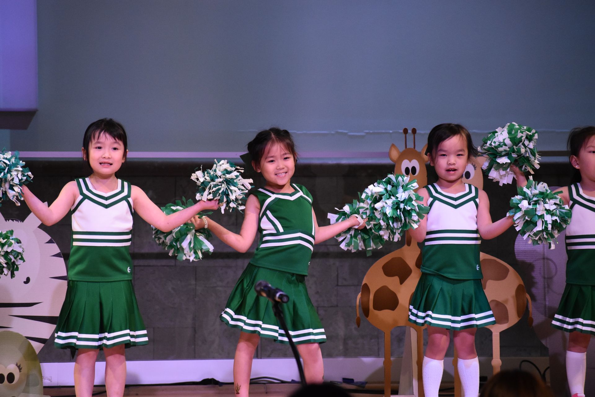 Children in green and white cheerleader outfits perform on stage with pom-poms.
