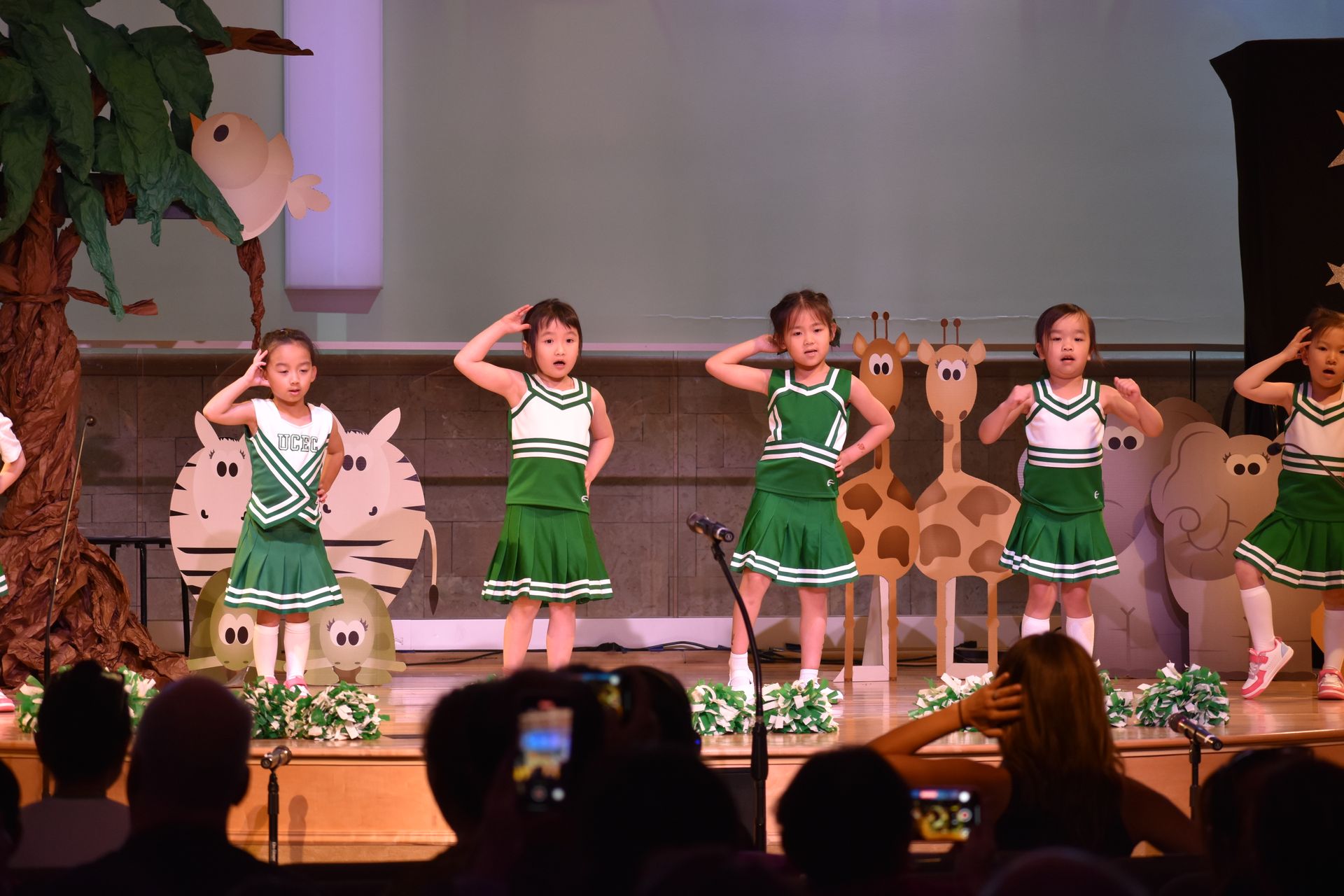 Five young performers in green and white outfits dance on a stage decorated with animal cutouts.