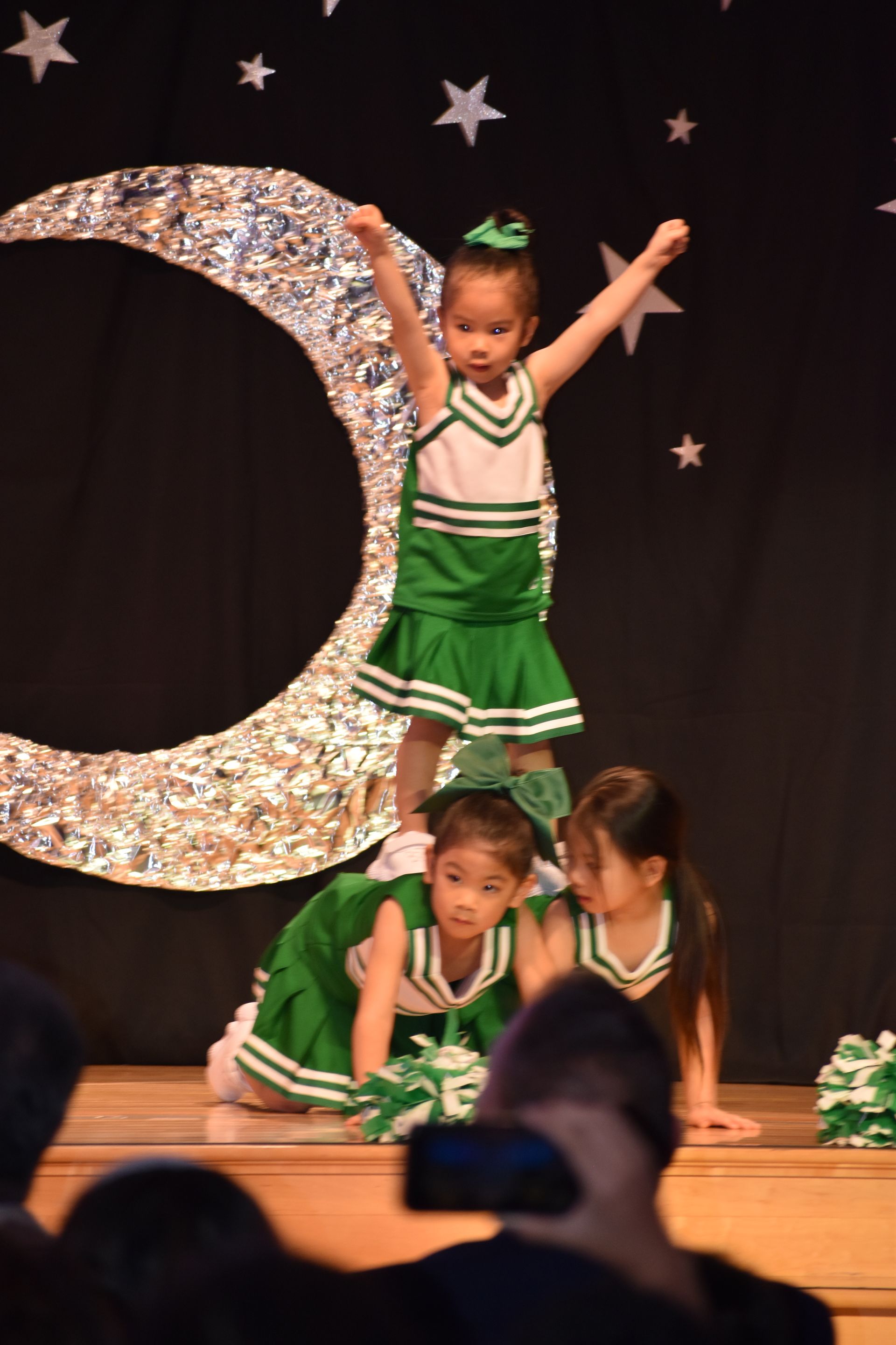 Young cheerleaders in green and white perform a pyramid on stage with a moon backdrop.