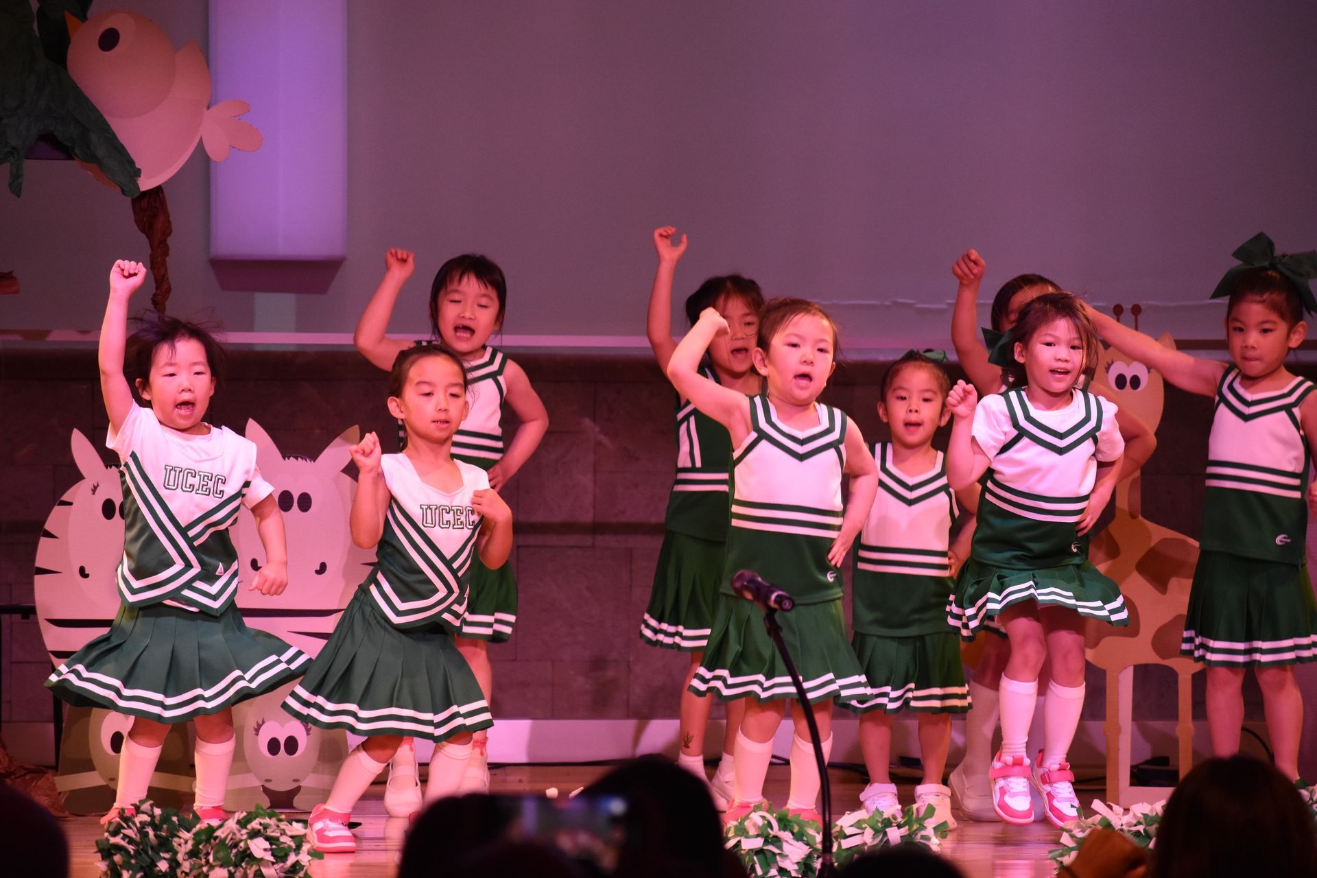 Children in green and white cheerleader outfits performing on a stage.
