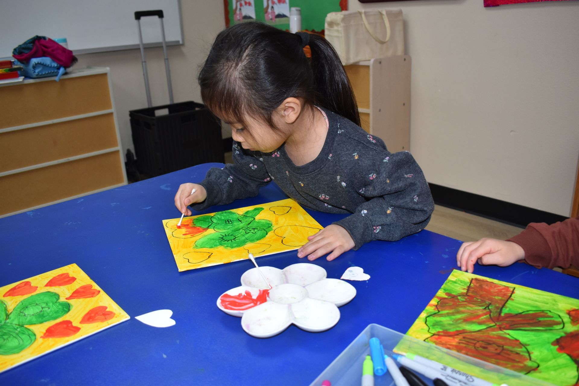Girl painting with paint on a yellow background at a blue table in a classroom.
